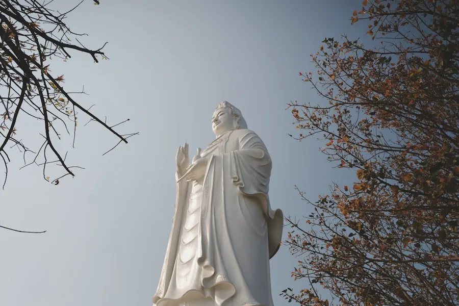 Ilustrasi mimpi kwan im — White statue of a deity framed by autumn trees.